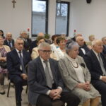 Grupa starszych osób w formalnych strojach siedzi na uroczystości w jasnym pomieszczeniu. W tle okna i ściana z krzyżem. Dekoracje stołów z owocami i świecami.