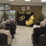 Spotkanie na sali z choinkami, złote dekoracje i napis Złote Gody na tle. Goście siedzą w rzędach. 행사 분위기 jest formalny.