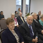 Grupa seniorów w eleganckich strojach siedzi w sali z białymi ścianami. Kolory dominujące to ciemne garnitury i czerwone ubrania. W tle widać okno i zastawiony stół.