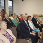 Grupa starszych osób siedzi na krzesłach w jasnym pomieszczeniu. Widoczne są eleganckie stroje: marynarki, białe bluzki. Na stole w tle stoją świeczniki i kwiaty.