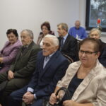 Grupa starszych osób siedzi w eleganckich strojach, na jasnym tle, z oknem i świecą w tle.