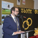 Mężczyzna w granatowym garniturze przemawia przy mikrofonie. W tle czarne tło z napisem ZŁOTE GODY i złotymi obręczami. Obok ozdoby kwiatowe i choinka.