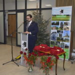 Na zdjęciu mężczyzna przy mównicy, za nim choinka i plakaty. Obrus jest bordowy, wazon z czerwonymi różami stoi obok. Tło jest jasne, widoczne flagi.