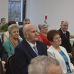 Grupa starszych osób siedzi w pomieszczeniu. Tło jest jasne z dekoracjami i świecami. Wszyscy są elegancko ubrani.