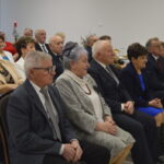 Grupa elegancko ubranych starszych osób siedzi na uroczystości. W tle biały stół z dekoracjami i czerwonymi świecami. Atmosfera oficjalna.