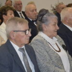 Grupa starszych osób siedzi w rzędzie w eleganckich strojach. Dominują odcienie szarości i niebieskiego. W tle widać elementy dekoracyjne.