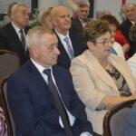 Grupa starszych osób siedzi na sali. Tło jest jasne, z czerwonymi świecami. Dominują kolory niebieski, beżowy i czarny.