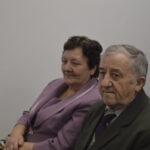 Nie znam tych osób, ale zdjęcie przedstawia dwie starsze osoby siedzące w pomieszczeniu o białych ścianach. Kobieta w liliowym kostiumie, mężczyzna w ciemnej marynarce. Obok widoczna inna osoba.