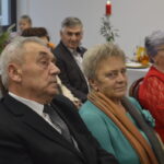 Grupa starszych osób siedzi przy stole na białym tle. Kobieta w niebieskim swetrze i szalu, mężczyzna w garniturze. Dekoracje świąteczne z tyłu.