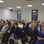 Ludzie w eleganckich ubraniach siedzą w jasnej sali, na ścianie krzyż, za oknami drzewa. Dominuje kolor granatowy i biały.