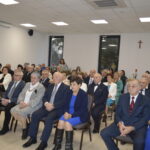 Grupa starszych osób siedzi w jasnej sali konferencyjnej. W tle białe ściany z krzyżem, okna z ciemnymi ramami. Osoby są elegancko ubrane.