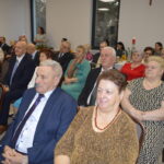 Grupa starszych osób siedzi na przyjęciu w eleganckiej sali. W tle widać duże okna i jasne ściany. Na stołach ustawione są świece i dekoracje.