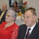 Zdjęcie przedstawia osoby siedzące w sali przy stole. Kobieta w czerwonej bluzce, mężczyzna w garniturze. W tle są inne osoby i zapalona świeca.