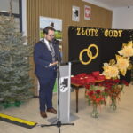 Mężczyzna w garniturze przemawia przy podium. Obok choinka z lampkami i dekoracje Złote Gody z kwiatami. Na stole czerwone pudełka i róże.