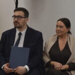 Para siedzi na ciemnoszarych krzesłach w jasnym pomieszczeniu. Obok choinka z lampkami. Kobieta i mężczyzna w eleganckich strojach.