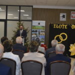 Sala z dekoracją w kolorach złotym i czarnym z napisem ZŁOTE GODY. Mężczyzna przemawia przy mikrofonie. W tle choinka i plakaty.