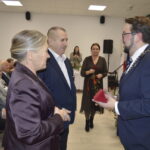 Sala z jasnym sufitem, ozdobiona choinką. Mężczyzna w garniturze wręcza pudełko parze starszych osób. W tle siedzi publiczność.