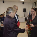 Na zdjęciu widać grupę osób w sali, kobieta wręcza różę. W tle choinka i biało-czerwone tła. Atmosfera oficjalna.