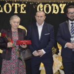 Na zdjęciu troje osób w eleganckich strojach. Starsza kobieta trzyma czerwoną różę, obok mężczyzna i urzędnik w łańcuchu. Tło czarne z napisem ZŁOTE GODY i światełkami.