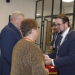 Grupa osób podczas ceremonii w eleganckiej sali, dominują kolory niebieski i złoty. Mężczyzna w garniturze wręcza medal kobiecie. W tle widoczny jest plakat z napisem Gmina Dobre.