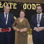 Na tle czarnej kurtyny z napisem „Złote Gody” stoją trzy osoby. Mężczyzna po lewej trzyma pudełko, kobieta w środku trzyma różę. Po prawej stronie jest mężczyzna z łańcuchem na szyi.