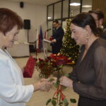 Dwie kobiety wymieniają uściski dłoni, jedna wręcza różę. W tle choinka z lampkami i mężczyzna przy mikrofonie.