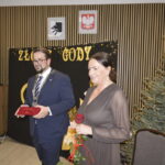 Na zdjęciu widać kobietę i mężczyznę w eleganckich strojach. Tło zdobi choinka i napis Złote Gody. Obok znajduje się baner promujący gminę.