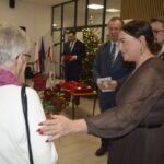 Na obrazie jest eleganckie spotkanie z choinką i flagami w tle. Kobieta w ciemnej sukni wręcza starszej pani różę. Obok stoją mężczyźni w garniturach.