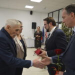 Na zdjęciu jest grupa osób uśmiechających się i podających sobie ręce. Ich ubrania są eleganckie, z dominacją ciemnych kolorów. W tle jest choinka ozdobiona światełkami.