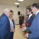 Starsza para w eleganckich ubraniach odbiera gratulacje i różę od mężczyzny w garniturze. W tle siedzą ludzie na krzesłach. Jasne wnętrze z dekoracjami.