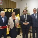 Cztery osoby w eleganckich strojach na tle dekoracji Złote Gody i choinki. Dwie osoby trzymają różę i czerwoną szkatułkę. Ściana ozdobiona światłami.