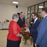 Grupa osób w eleganckich strojach, starsza kobieta w czerwieni otrzymuje kwiaty w jasnym pomieszczeniu. Tło: stół z białym obrusem, dekoracje, choinka. Klimat uroczysty, oficjalny.