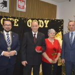 Na zdjęciu cztery osoby stojące przed czarnym tłem z napisem Złote Gody. Mężczyzna trzyma czerwoną kopertę, kobieta czerwoną różę. Obok nich choinka ozdobiona światełkami.