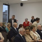 Grupa starszych osób siedzi na uroczystości w jadalni z dużymi oknami w tle. Wszyscy ubrani elegancko, atmosfera formalna. W tle widoczne są czerwone świece i dekoracje.