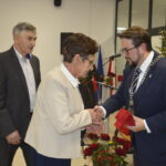 Na obrazie widoczne są trzy osoby w eleganckiej sali. Mężczyzna w garniturze wręcza starszej kobiecie prezent. W tle choinka i dekoracje świąteczne.