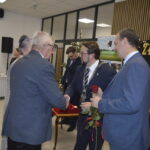 Mężczyzna w garniturze ściska rękę starszemu panu w sali bankietowej. W tle stół z białym obrusem i dekoracje. Dominują odcienie szarości i niebieskiego.