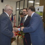 Na zdjęciu widoczne są trzy osoby ubrane formalnie, przekazujące sobie czerwone róże. W tle sala z flagami i białymi ścianami. Złoty napis Złote na ciemnym tle po prawej stronie.