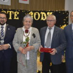 Cztery osoby stoją przed czarnym tłem z napisem ZŁOTE GODY. Starsza kobieta trzyma różę, a obok niej mężczyzna z pudełkiem. W tle choinka i światełka.