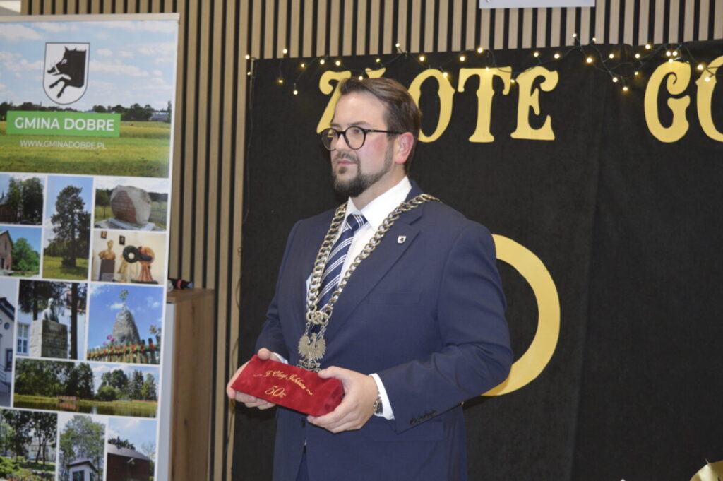 Na zdjęciu widać mężczyznę w garniturze z łańcuchem, trzymającego czerwony aksamitny worek. Tło jest czarne z napisem „ZŁOTE”. Obok stoi banner promujący Gminę Dobre.