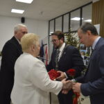 Na obrazie widać osobę starszą w białym ubraniu, która rozmawia z mężczyzną w garniturze. W tle znajduje się choinka z lampkami i stół z przekąskami. Całość ma formalny charakter, z delikatnym oświetleniem.