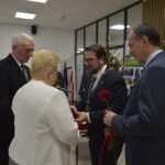 Grupa ludzi podczas uroczystości; mężczyzna z łańcuchem wręcza róże starszej parze. Tło zdobi choinka i złote napisy. Kolory dominujące to czarny, biały, czerwony.