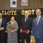 Cztery osoby w eleganckich strojach stoją przed tablicą z napisem „Złote Gody”. Tło czarne z żółtymi literami i lampeczkami. Jedna osoba trzyma różę, inna pudełko.