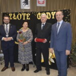 Cztery osoby w eleganckiej odzieży stoją przed czarną ścianką z napisem Złote Gody. Kobieta trzyma bukiet róż, a mężczyzna pudełko. W tle znajduje się choinka z lampkami i baner z krajobrazami.