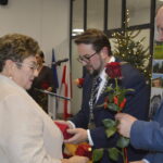 Na zdjęciu znajduje się mężczyzna wręczający kobiecie czerwoną różę. Tło stanowi choinka z lampkami i flagi w biurze. Atmosfera formalna, eleganckie ubrania.