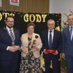Grupa osób w eleganckich strojach pozuje na tle czarnych złotych ozdób. Kobieta trzyma różę. W tle jest choinka.