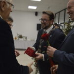 Grupa elegancko ubranych osób w garniturach rozmawia, trzymając róże. W tle jasne pomieszczenie, stół z obrusami i choinka. Atmosfera jest świąteczna i uroczysta.