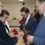 Na zdjęciu grupa osób wymieniających się czerwonym kwiatem i pudełkiem. Tło jasne, z białym stołem i dekoracjami. Ubrani formalnie w garnitury i eleganckie stroje.