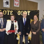 Grupa pięciu osób w eleganckich strojach na tle napisu Złote Gody. Kobieta trzyma różę i czerwoną szkatułkę. W tle choinka i ozdoby.