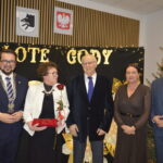 Pięć osób stoi przed czarnym tłem z napisami Złote Gody. Osoby są elegancko ubrane, jedna trzyma czerwoną różę i pudełko. W tle znajduje się choinka oraz godła.