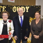 Na zdjęciu pięć osób pozujących podczas uroczystości. Tło jest ciemne z jasnymi napisami i ozdobami. Kobieta trzyma różę i czerwone pudełko.